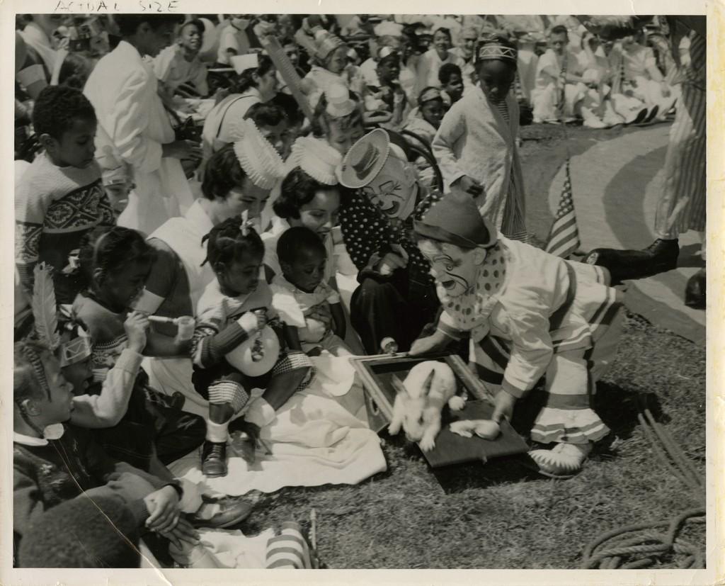 Two nurses sit with children in front of a pair of clowns during Circus Day, c. 1954