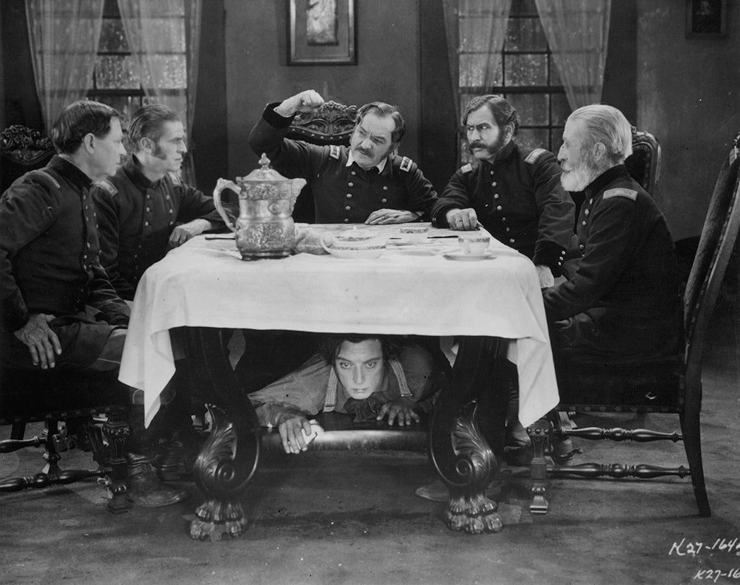 Buster Keaton hiding under table as group of men plan in a scene from the film 'The General', 1926