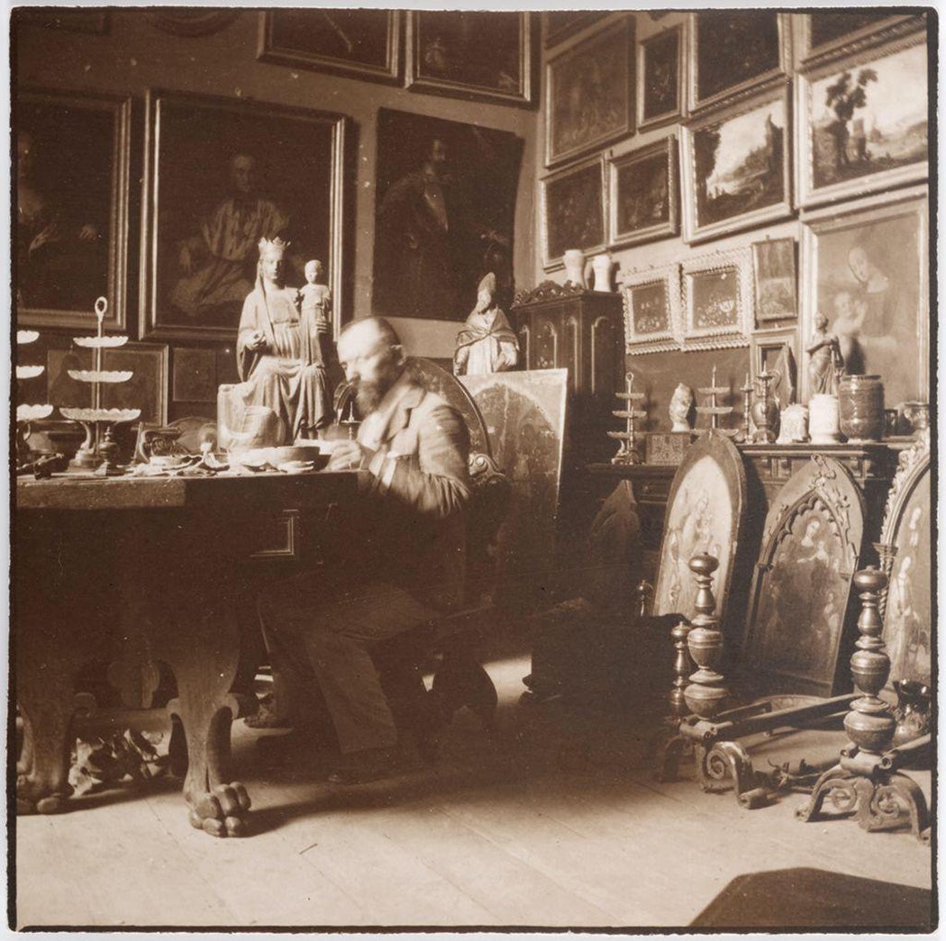 Bernard Berenson sitting at a table in a room filled with paintings.